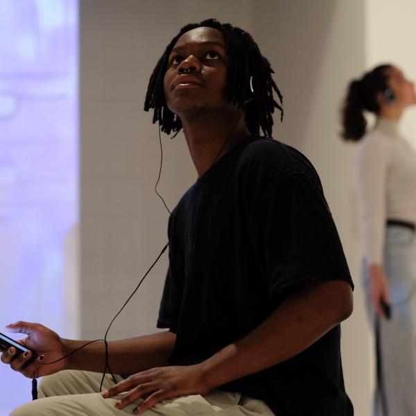 A young man wearing headphones and holding a mobile phone is sitting, and there's a young woman standing in the background, also wearing headphones and holding a phone..