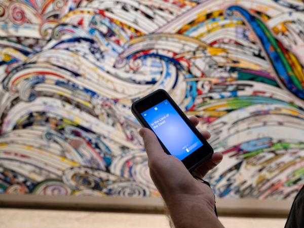 A hand holding an iPod Touch in front of a mural at Japan Supernatural exhibition at Art Gallery of New South Wales. The device shows an audio track is playing.