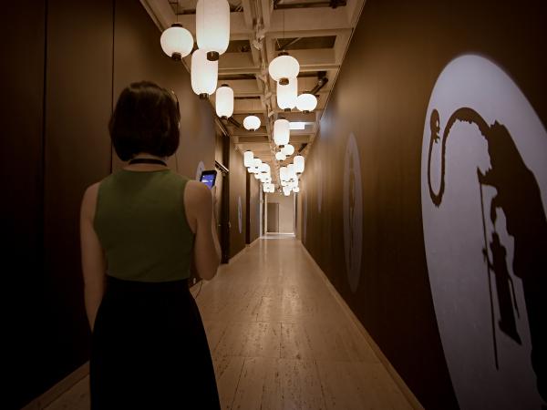 A woman wearing headphones is walking down a hallway at the Japan Supernatural exhibition at Art Gallery of NSW. She is looking at a iPod Touch in her hand, and artwork is projected onto the walls.