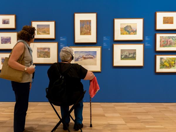 Two women wearing headphones looking at a wall of paintings. One woman has a mobility aid.