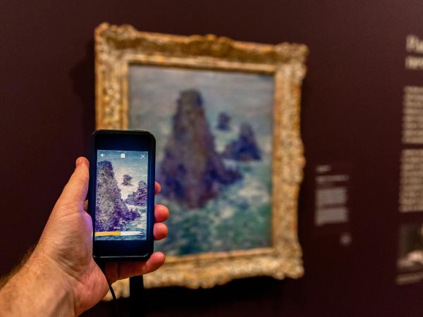 A hand holding an iPod Touch in front of a wall painting at the Josh Russell exhibition at the Art Gallery of NSW. The device shows the same painting on its screen.