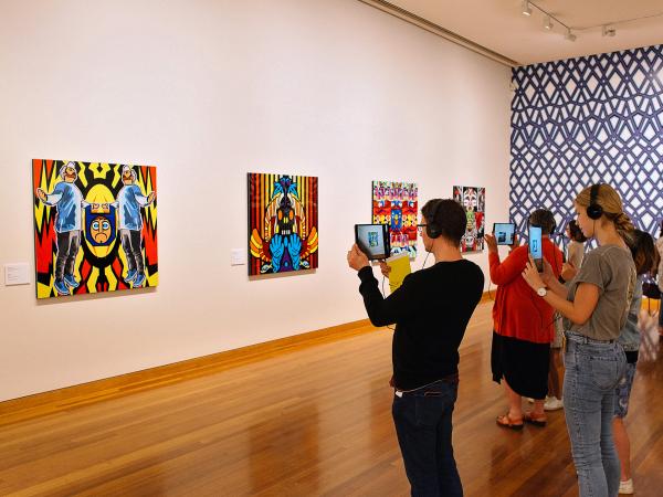 A group of people are wearing headphones and holding iPads up to Indigenous Artist John Muir's artwork to trigger an augmented reality experience at the Bendigo Art Gallery.