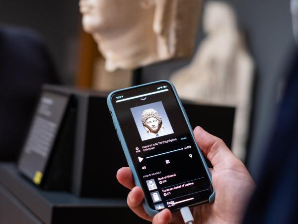 Close up photo of a man's hand holding a mobile phone showing the audio guide. There is a sculpture in the background