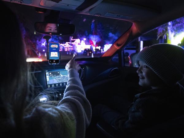 A mother and young son are sitting inside a car at the Elf on the Shelf Magical Holiday Journey. There is a phone in a hands-free holder showing the Elf Attractions' location-aware UI and the mother is pointing to the Christmas displays outside