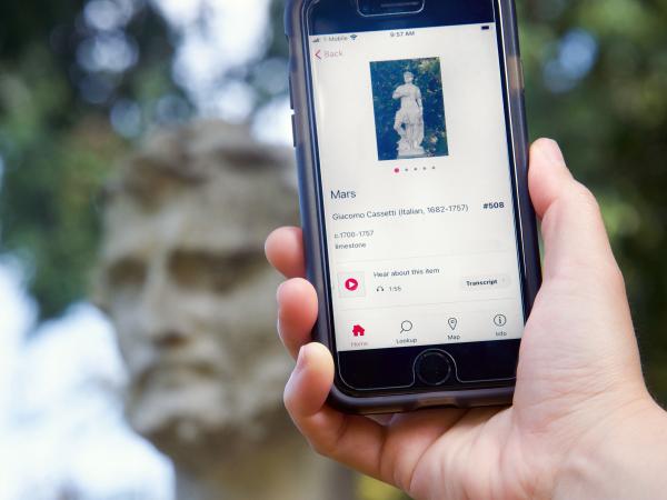 A hand holds a phone displaying information about the statue Mar, which is blurred in the background.