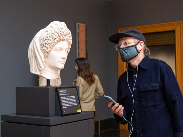 Man wearing a hat, mask and earphones and holding a mobile device looks at a bust inside the J. Paul Getty Museum.
