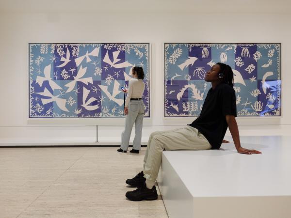 A young man wearing headphones sits and listens to the Matisse: Lift and Spirit audio guide, and there's a young woman standing in the background looking at one of Matisse's paintings.