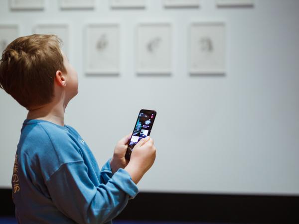 A child is looking up at wall art at the Museum of Old and New Art. He is holding a mobile device in his hand that is showing the interface from The O application.