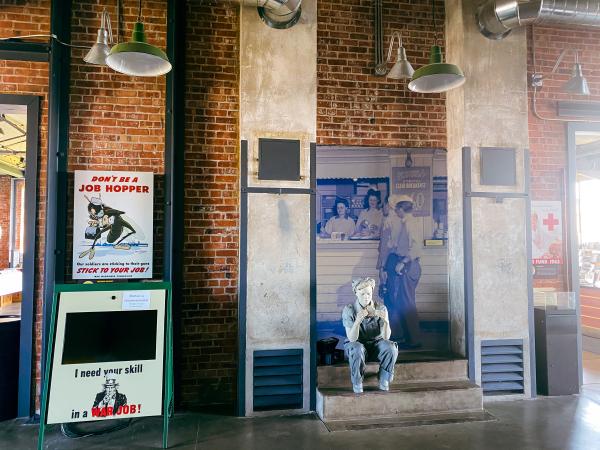 Display at the Rosie the Riveter exhibition for the National Park Service. A mannequin dressed as Rosie the Riveter sits on a step in front of historic memorabilia