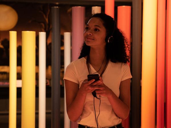 Young woman holding a mobile device and wearing headphones in a space with colourful poles behind her.