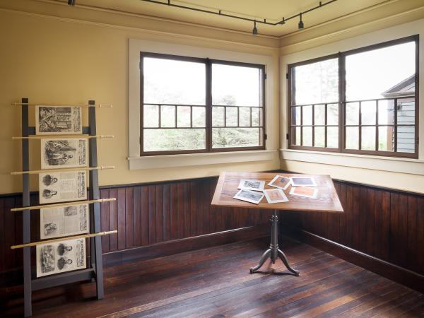 Inside Winslow Homer's studio, a magazine rack sits to the left with hanging newspapers and to the right a table with images.