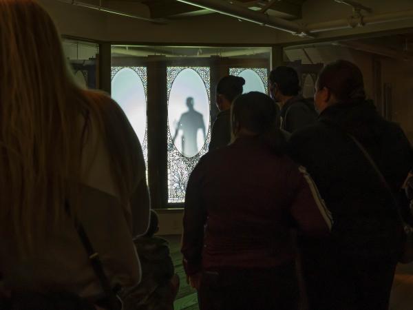 A ghostly image stands behind a glass inlaid door, projected onto the glass as people look on with their backs in view.