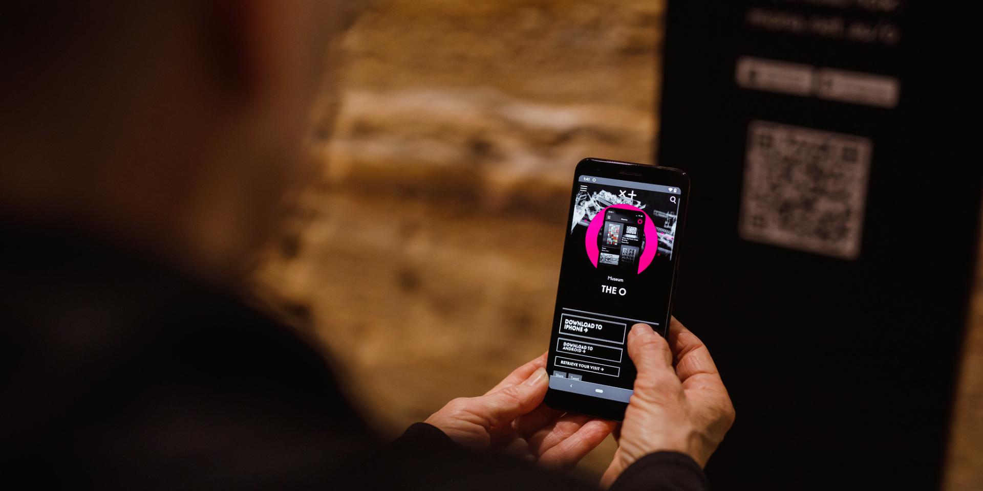 Person holding a mobile device with Mona's The O displaying on the screen.