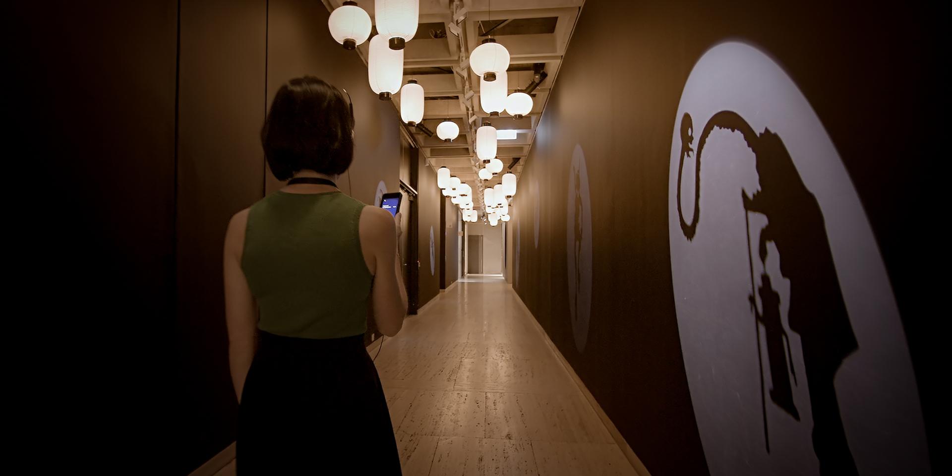 A woman wearing headphones is walking down a hallway at the Japan Supernatural exhibition at Art Gallery of NSW. She is looking at a iPod Touch in her hand, and artwork is projected onto the walls.