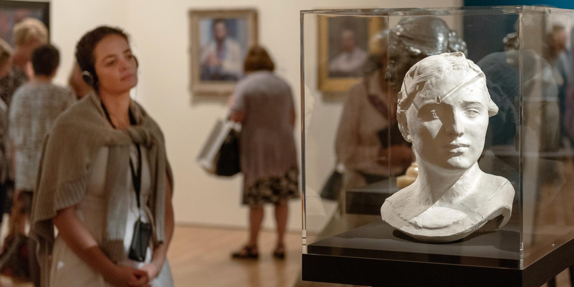 A woman wearing headphones is looking at a sculpture of a bust at the Josh Russell exhibition at the Art Gallery of NSW. She has an iPod Touch device on a lanyard and is listening to an audio guide