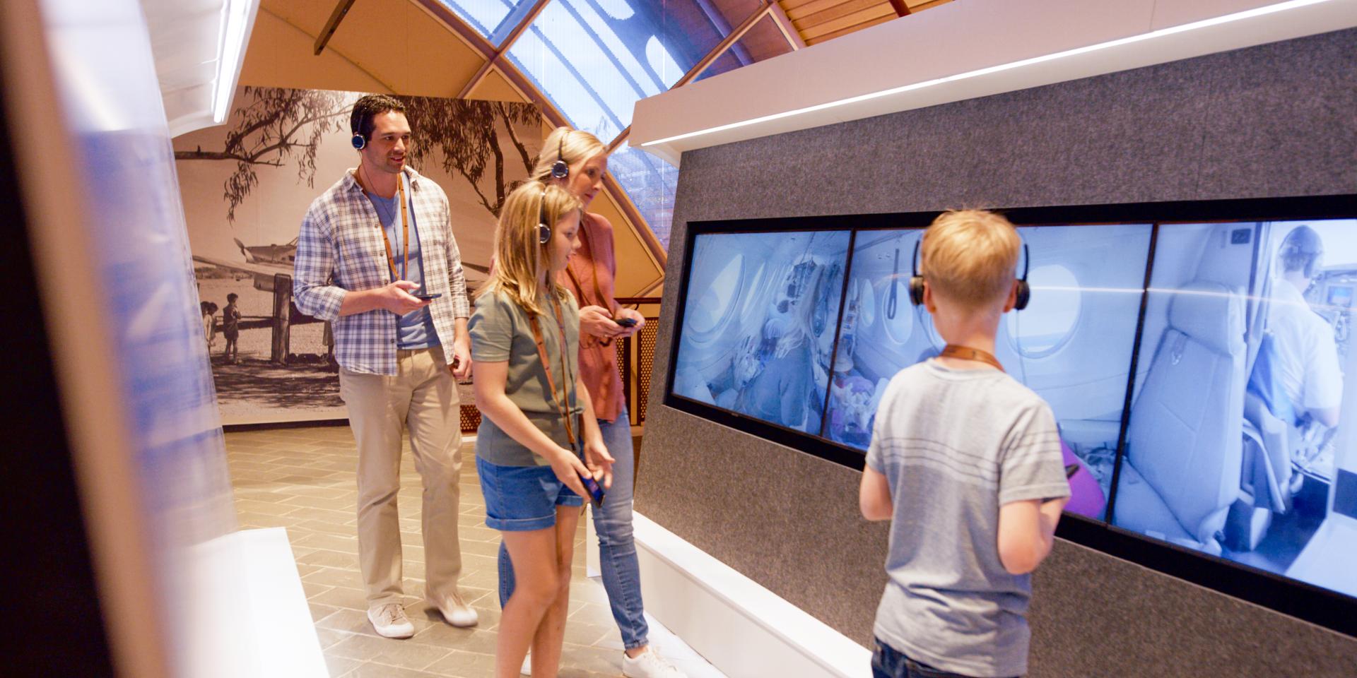 Two adults and two children walk past video screens at the Australian Stockmans Hall of Fame. They are all wearing headphones and listening to an audio guide