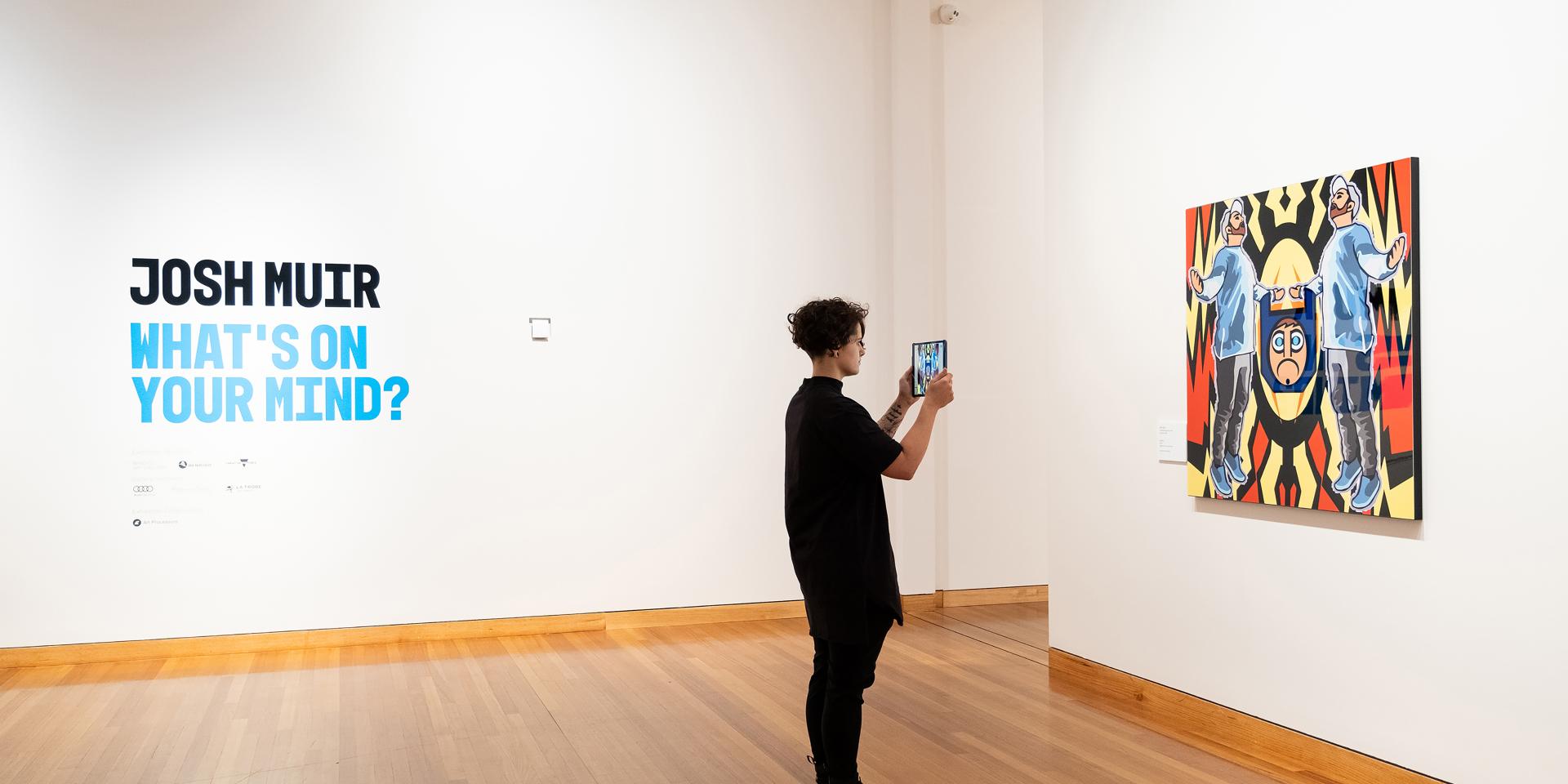 A woman stands in front of Josh Muir's artwork at the Bendigo Art Gallery. She is holding an iPad up to the artwork to trigger an augmented reality experience