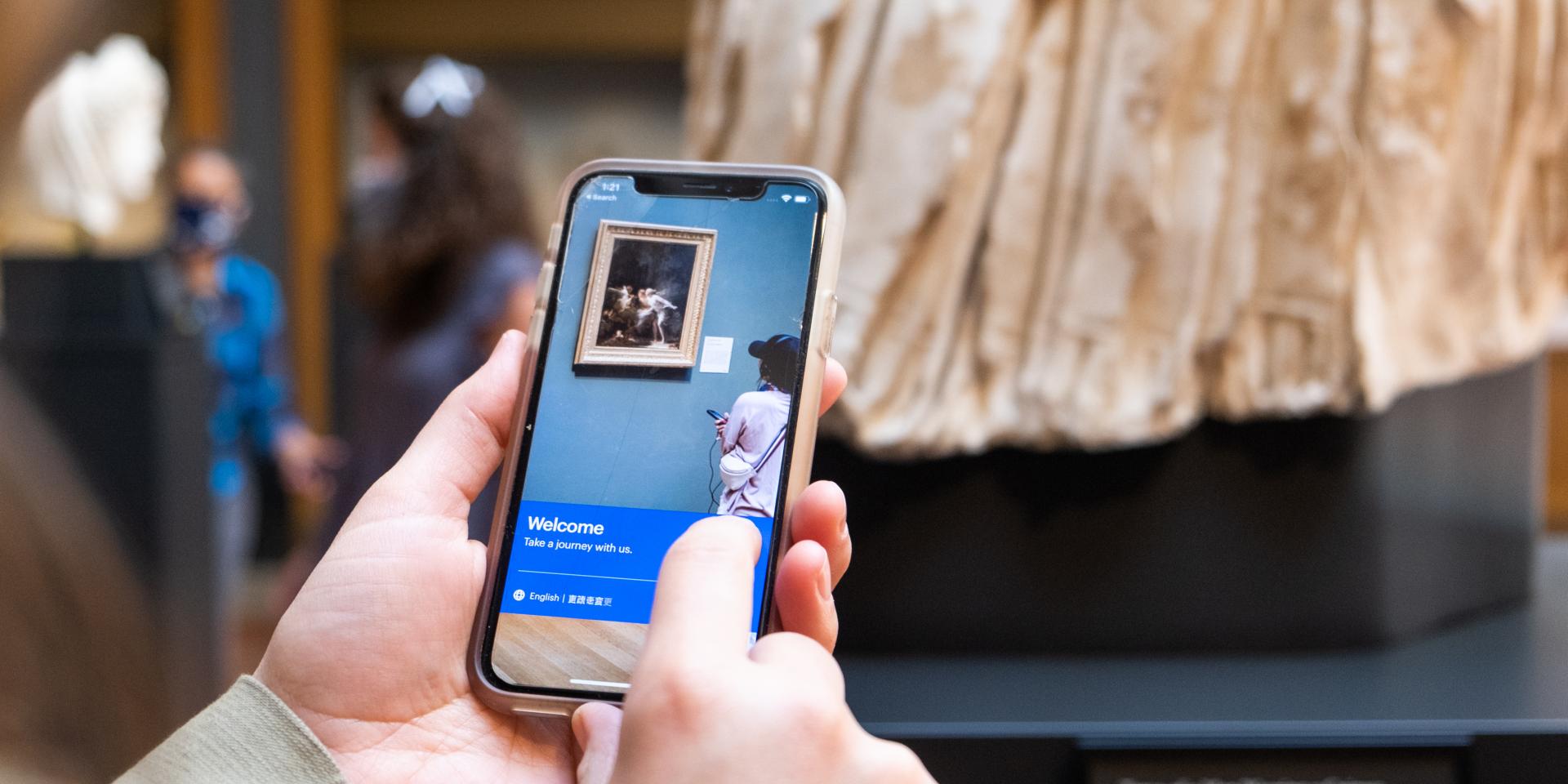Close up photo of a man's hand holding a mobile phone showing the audio guide. There is a sculpture in the background