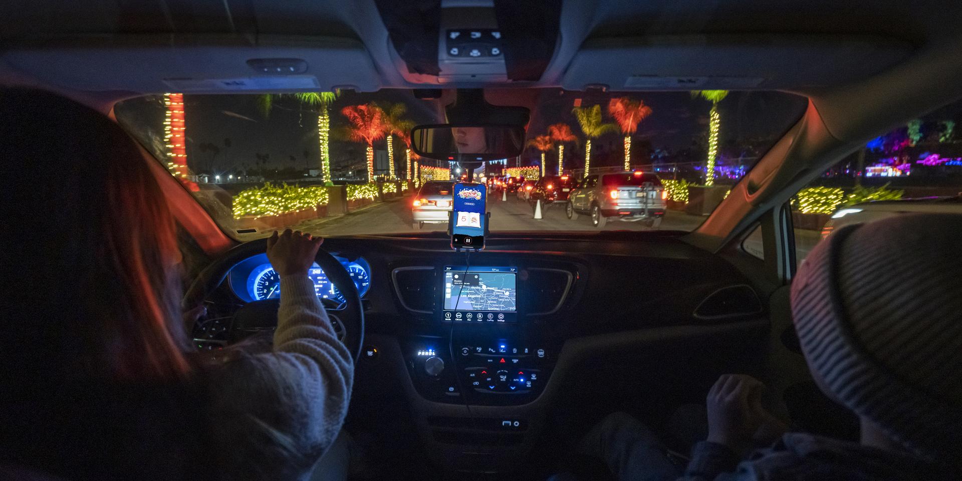 A mother and young son are sitting inside a car at the Elf on the Shelf Magical Holiday Journey. There is a phone in a hands-free holder showing the Elf Attractions' location-aware UI