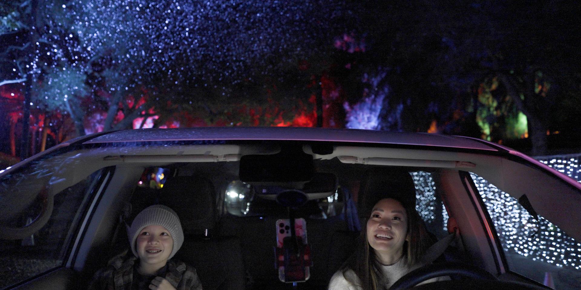 Mother and young son sitting in their car at night during the Elf on the Shelf Magical Holiday Journey. Their faces show wonder and are surrounded by Christmas lights