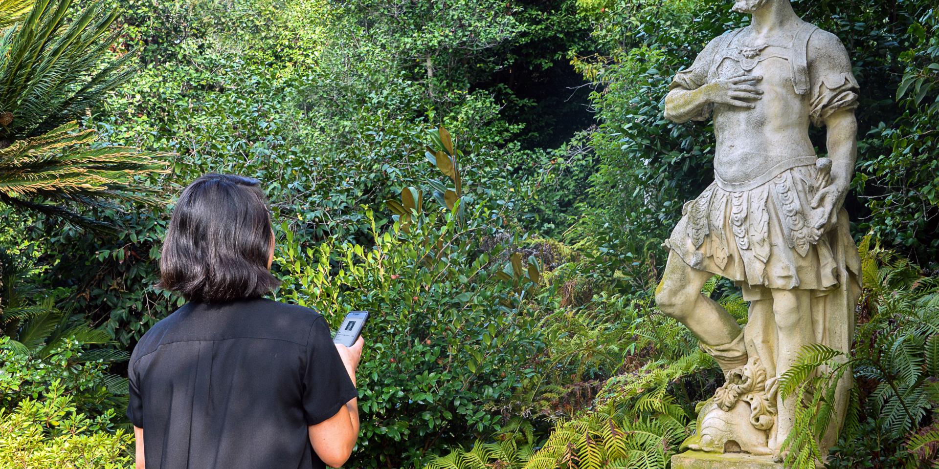 A women wearing a black top and holding a phone is facing a Roman statue.