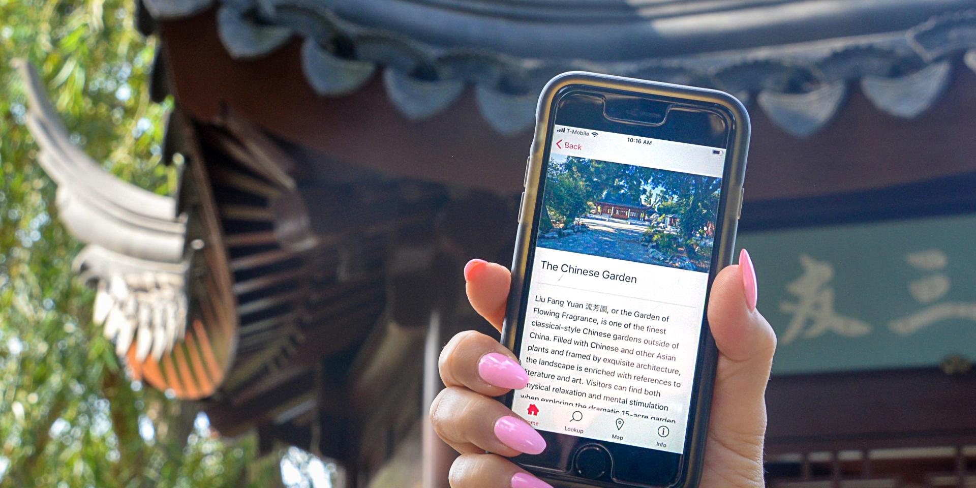 A hand with fingernails painted pink holds a mobile phone in front of a building with a decorative Chinese roof.