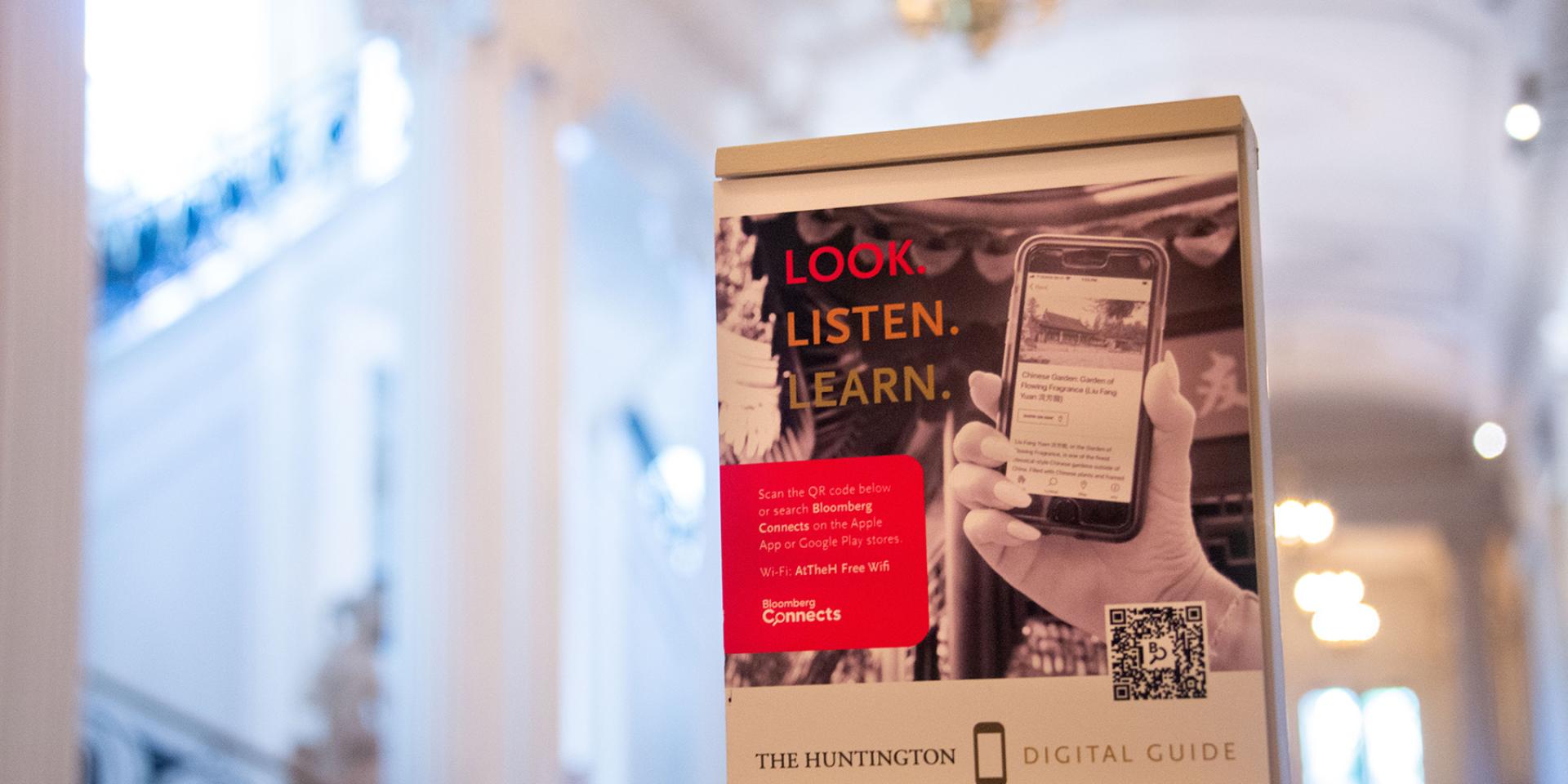 A sign inside the The Huntington promoting the center's audio guide, showing a hand holding a mobile phone.