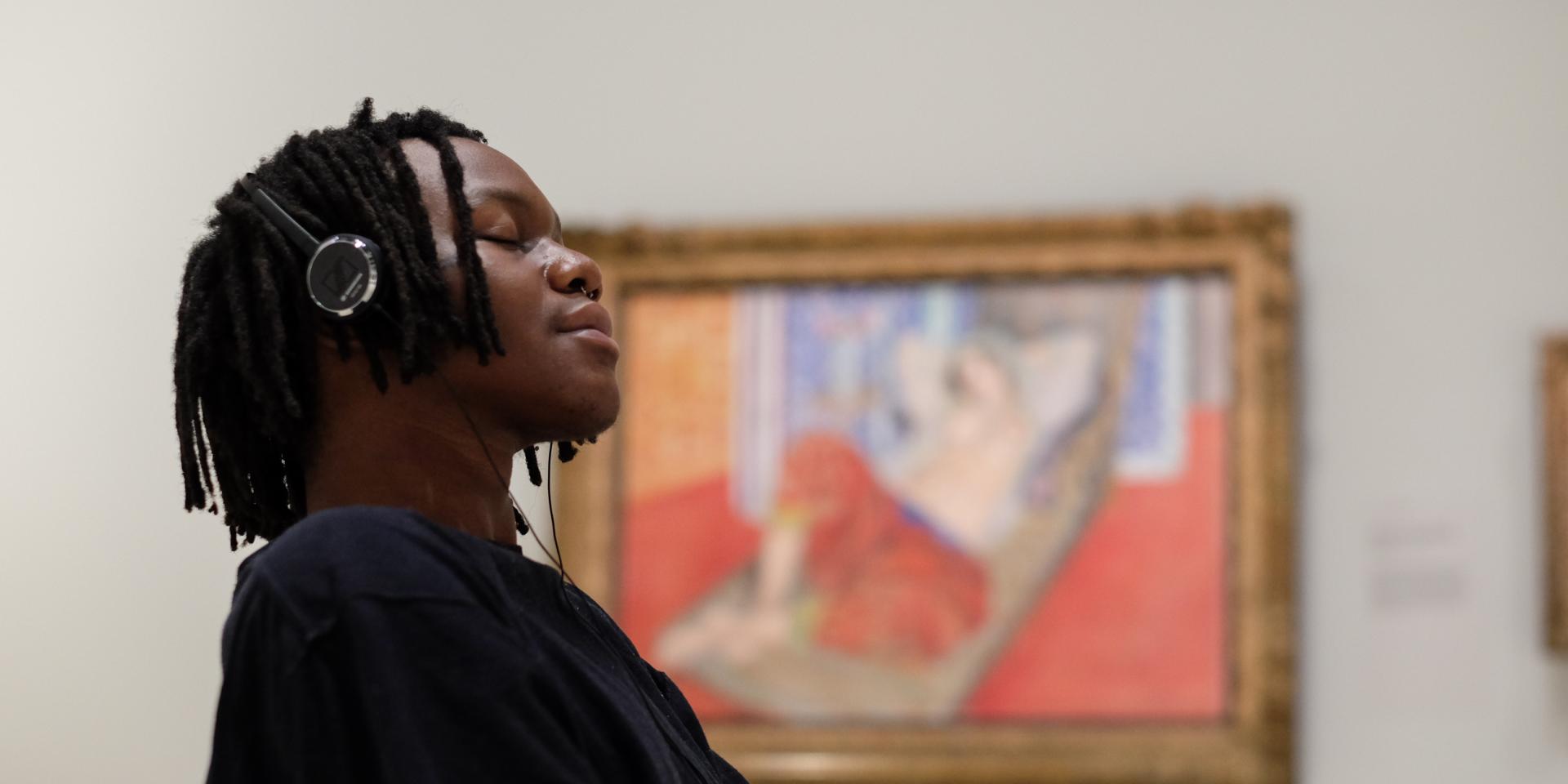 A young man wearing headphones sits and listens to music from the Matisse: Life and Spirit audio guide, with a painting by Matisse in the background.