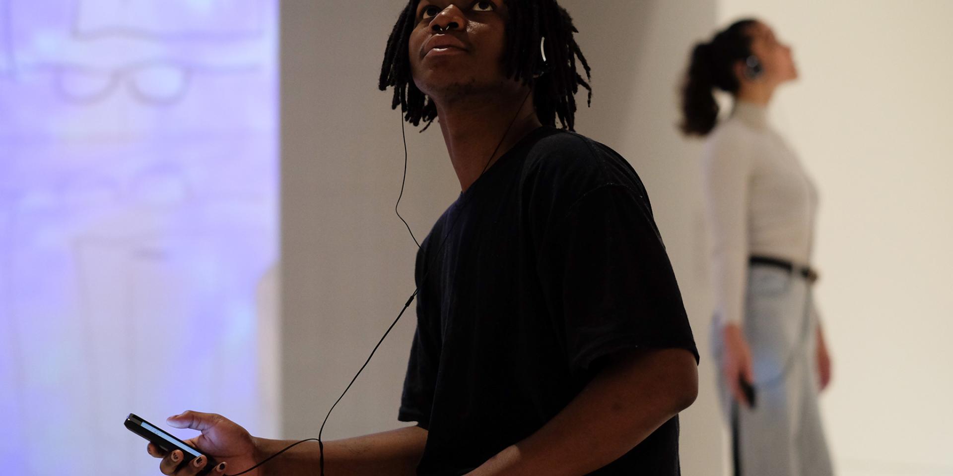 A man wearing a black t-shirt who is sitting in a gallery is looking up and holding a mobile phone and wearing headphones. In the background is a woman wearing jeans and a white top, also looking up and holding a mobile phone and wearing headphones.