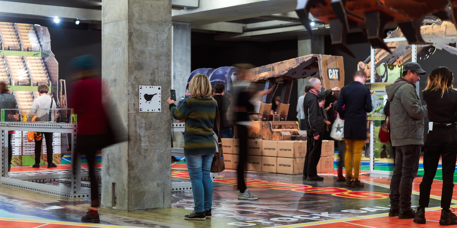 A woman stands in front of an augmented reality marker holding up her phone at the Simon Denny:Mine exhibition at Mona