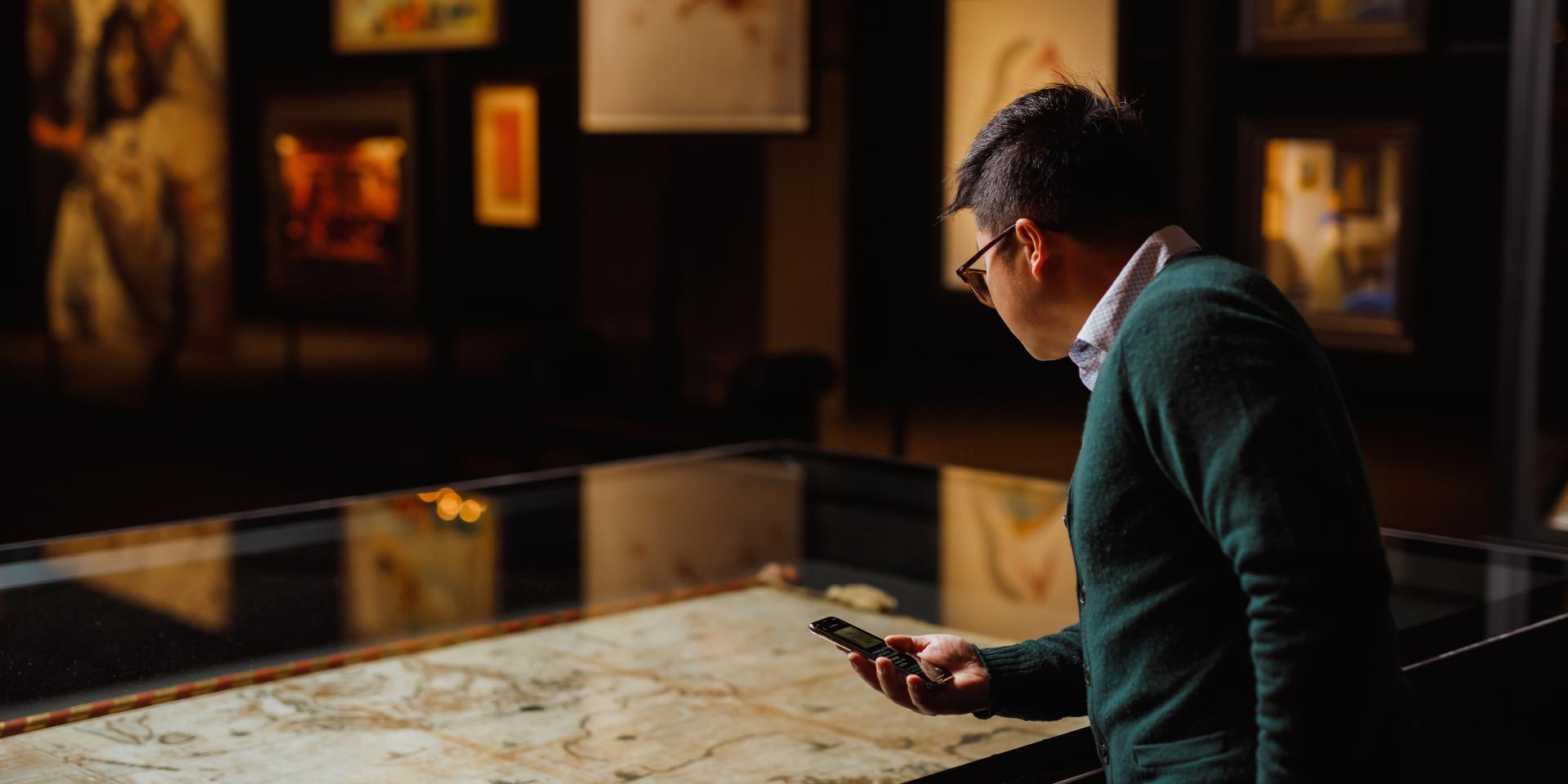 A man standing in front of an artwork display case. He is holding a mobile device in front of him