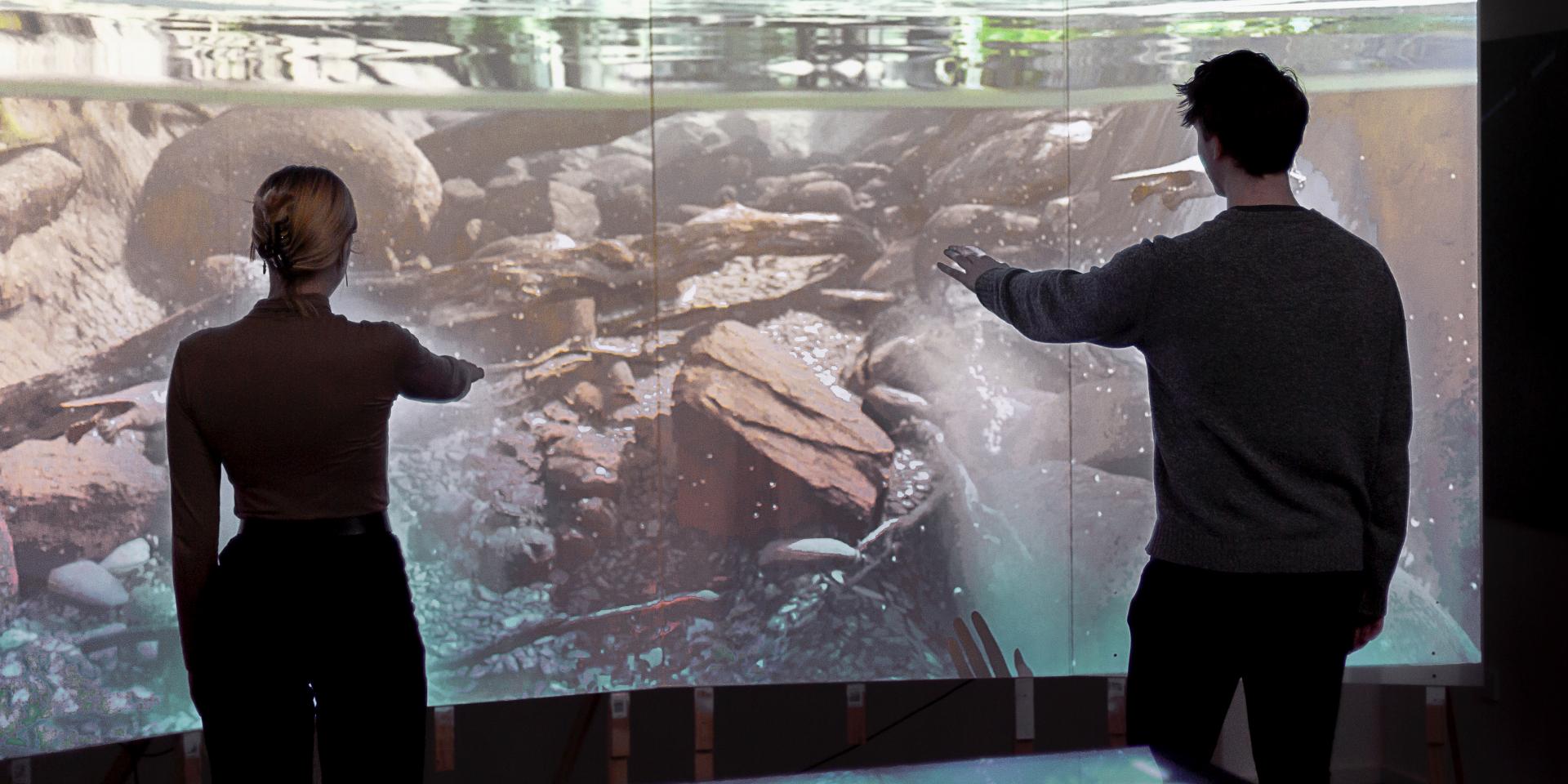 A woman and man stand in front of a projection screen. They both have their arms lifted and are interacting with the projection which tracks their movement 