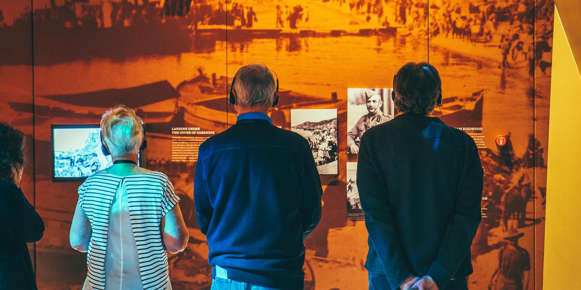 A group of people stand in front of a display wall at the Spirit of the ANZAC touring exhibition. They are all wearing headphones and listening to an audio guide