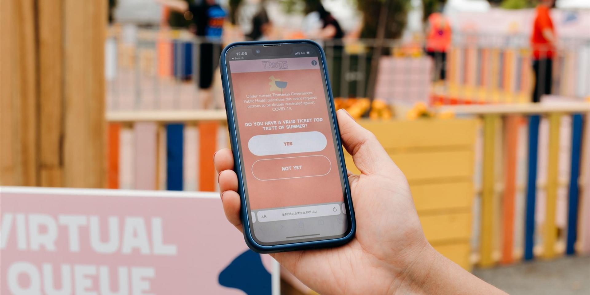 A hand holding a phone displaying the Taste of Summer virtual queuing web app.