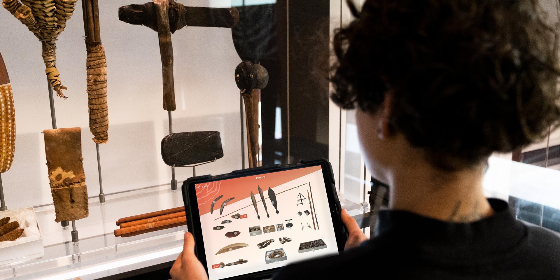 A woman is holding an iPad at the Awaken exhibition. The iPad content replicates the objects in a showcase in front of her