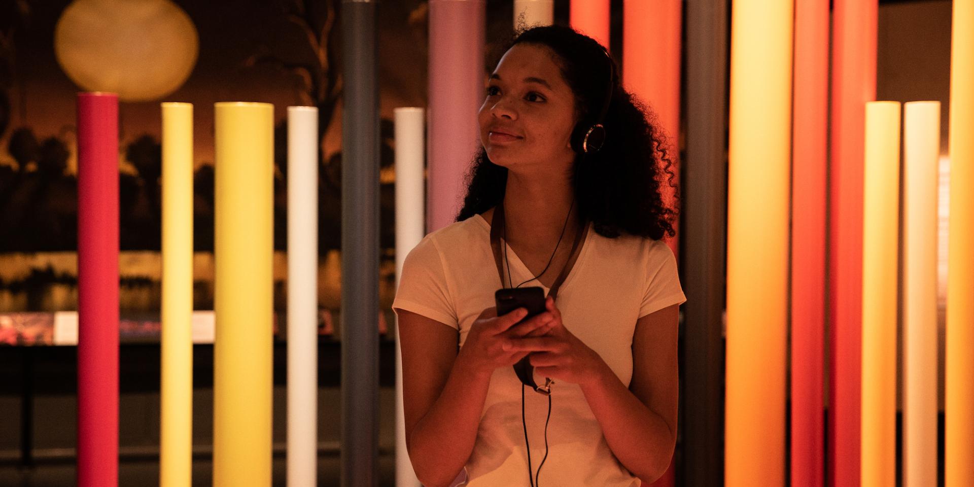 Young woman holding a mobile device and wearing headphones in a space with colourful poles behind her.