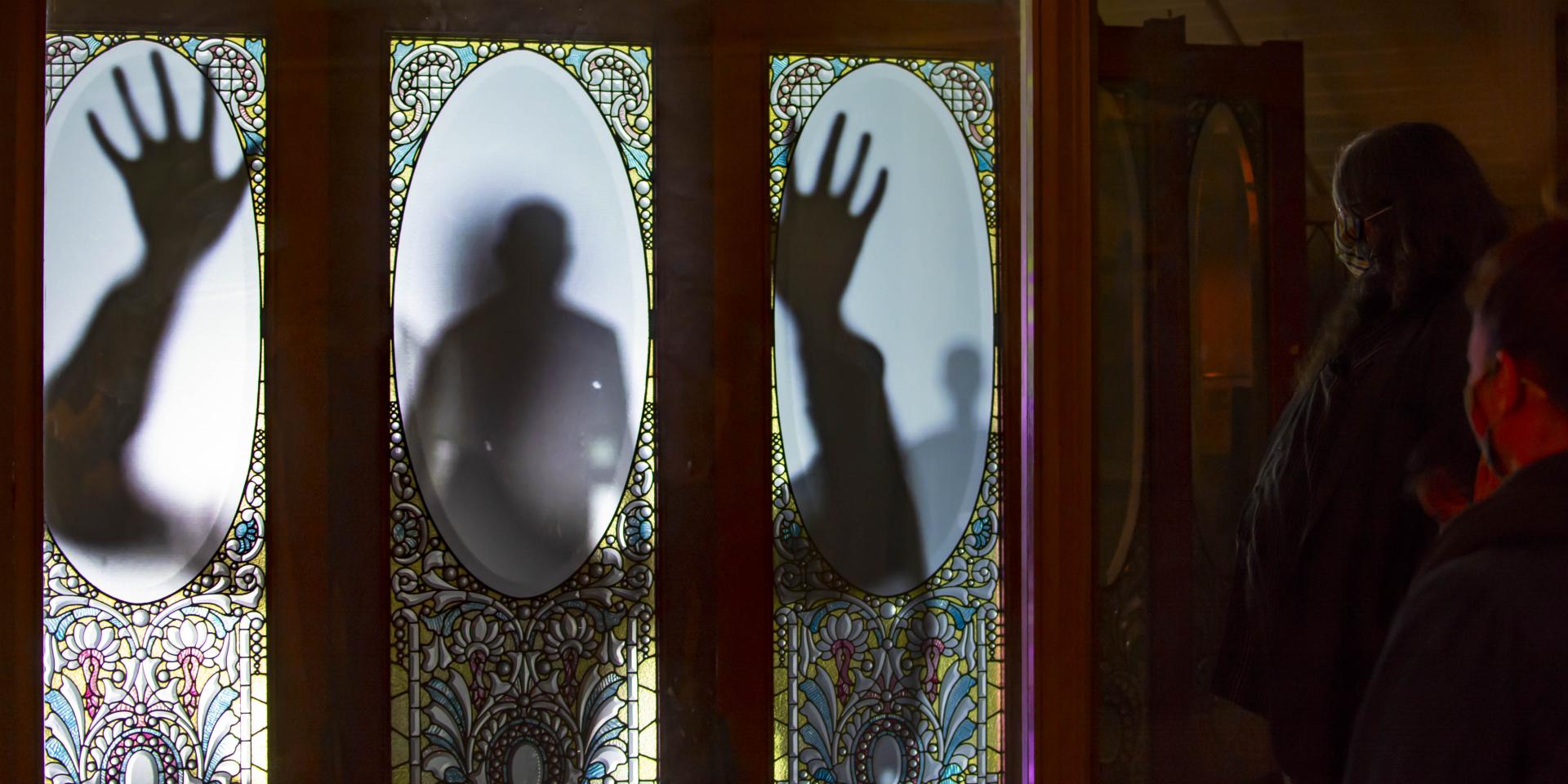 The projected image of person leaning against a glass inlaid door, as two visitors to Winchester Mystery House look on.