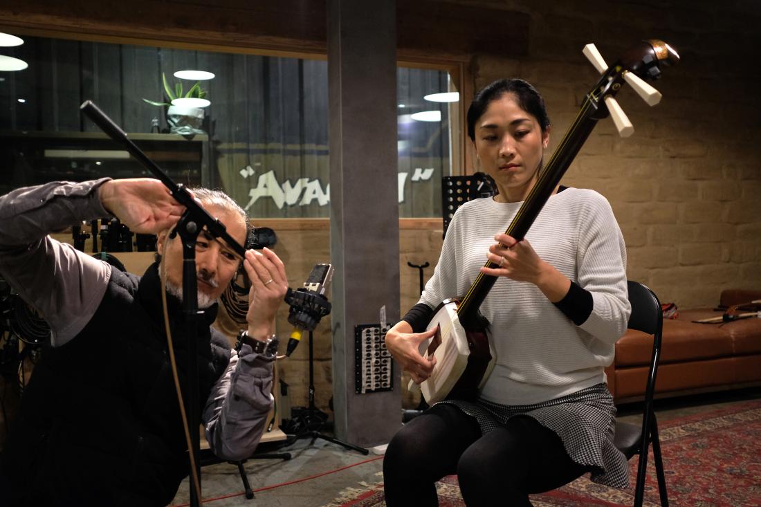 A man and woman are in a sound recording studio. He is adjusting a microphone and she is playing a Japanese shamisen.