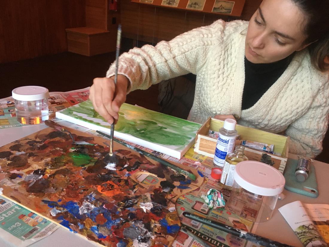 A woman holding a paint brush with paint suppliers all over a table.