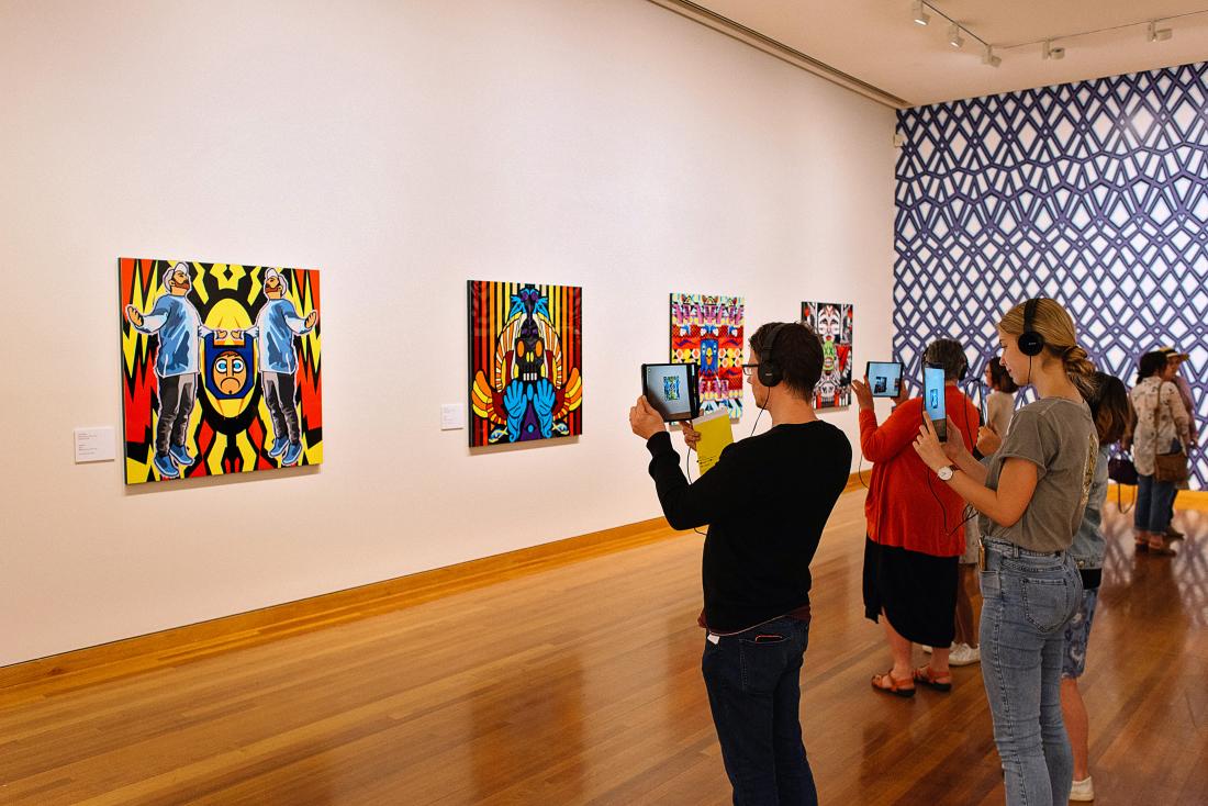 A group of people are wearing headphones and holding iPads up to Indigenous Artist John Muir's artwork to trigger an augmented reality experience at the Bendigo Art Gallery.