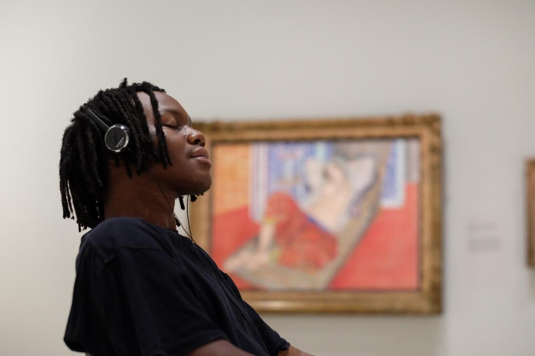 A young man wearing headphones sits and listens to music from the Matisse: Life and Spirit audio guide, with a painting by Matisse in the background.