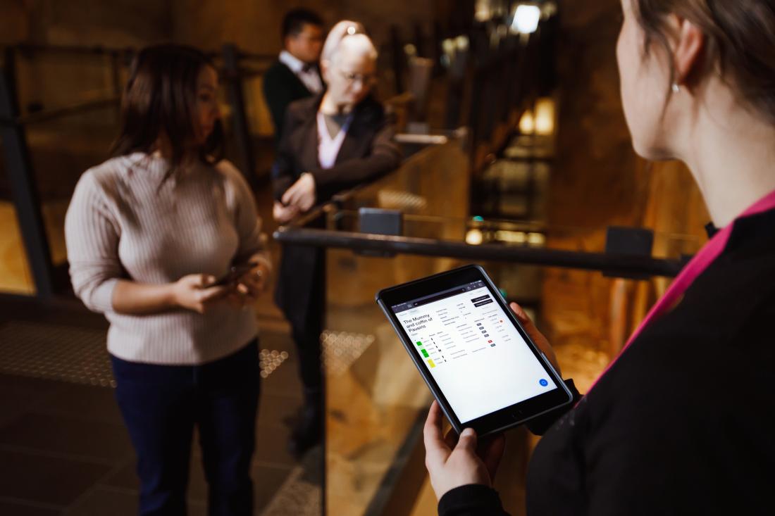 A Mona staff member is holding an iPad with The O's queuing technology displayed. She is standing in front of three waiting visitors