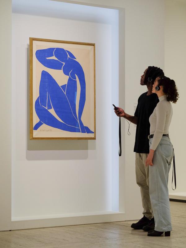 A wearing a black t-shirt and a woman wearing a white top and blue jeans are holding mobile phones and wearing headsphones while looking at a painting by Henri Matisse.