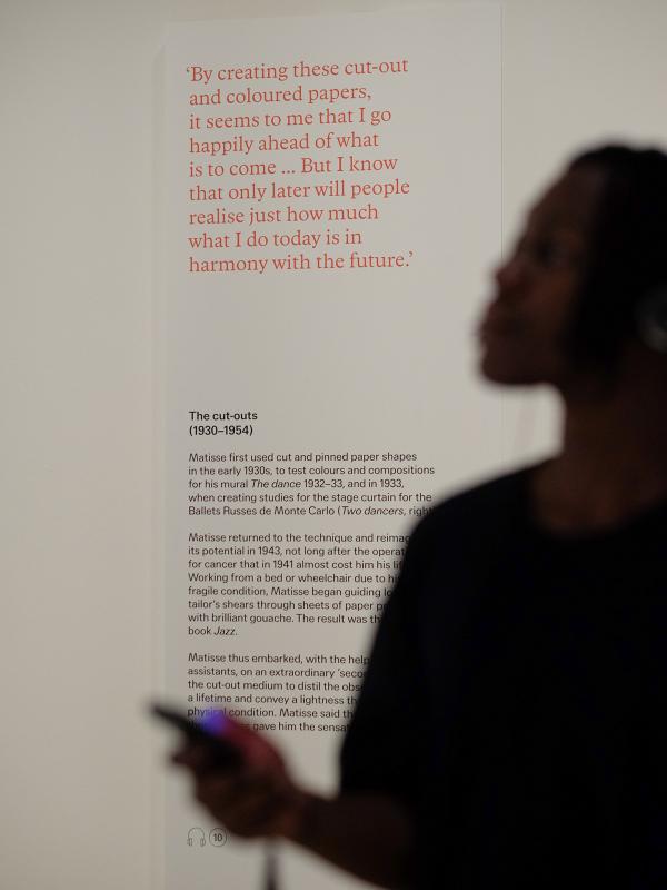In the background there's some text displayed on the wall of an exhibition about artist Henri Matisse's work. In the foreground there's a silhouette of a man wearing a black shirt and holding a mobile phone.